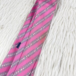 May include: A bright pink necktie featuring a repeating diagonal pattern of interlocking silver and white chain links. The fabric has a smooth, satin-like finish, and a small dark blue care tag is visible on the inner fold of the tie.