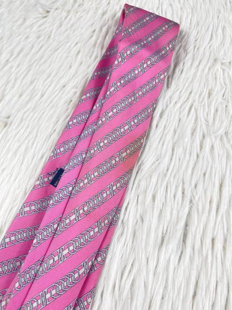 May include: A bright pink necktie featuring a repeating diagonal pattern of interlocking silver and white chain links. The fabric has a smooth, satin-like finish, and a small dark blue care tag is visible on the inner fold of the tie.