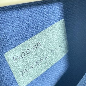 May include: A close-up shot of a blue fabric with a tag attached. The tag has handwritten text that reads "A500 HO" and "24 x 26 x 23.5". The fabric is a solid blue color and appears to be a knit material. A white hanger is visible at the top.