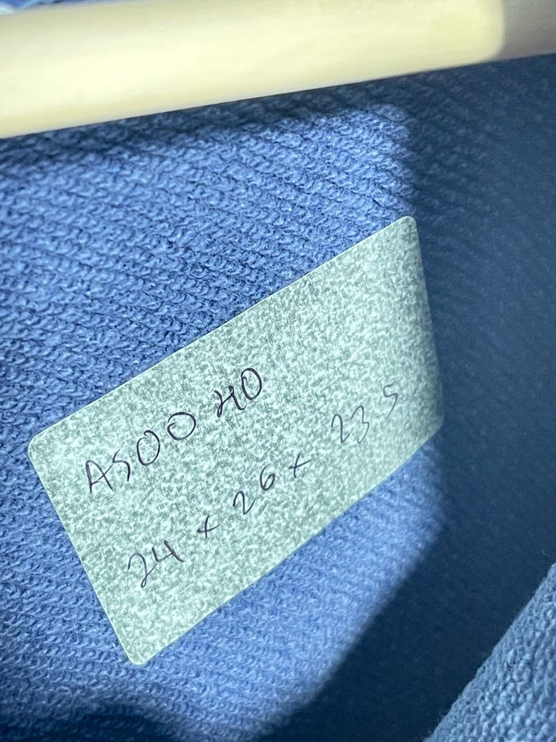 May include: A close-up shot of a blue fabric with a tag attached. The tag has handwritten text that reads "A500 HO" and "24 x 26 x 23.5". The fabric is a solid blue color and appears to be a knit material. A white hanger is visible at the top.
