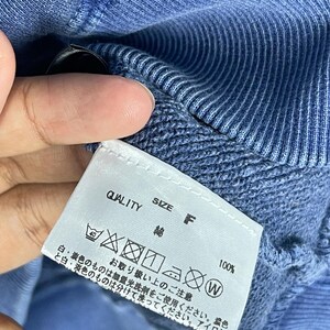 May include: Close-up of a blue garment with a white tag. The tag reads "QUALITY", "SIZE F", and includes washing instructions. The fabric appears to be cotton. The garment is a solid blue color.