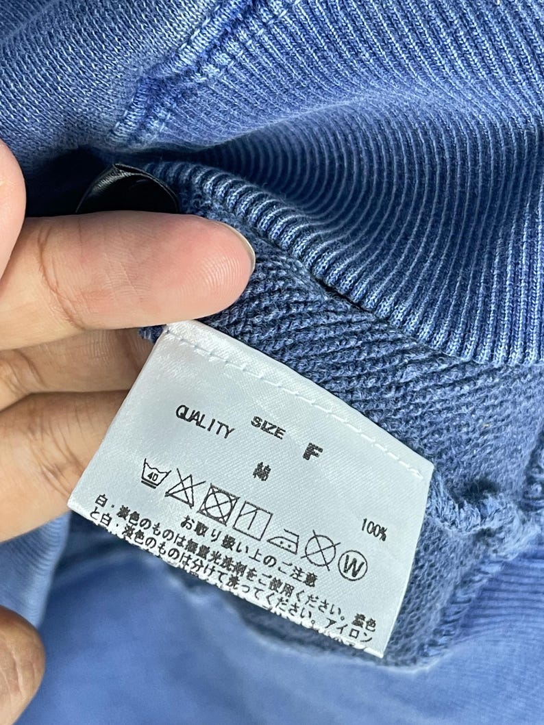 May include: Close-up of a blue garment with a white tag. The tag reads "QUALITY", "SIZE F", and includes washing instructions. The fabric appears to be cotton. The garment is a solid blue color.