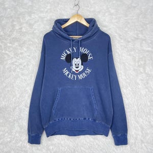 May include: A blue hooded sweatshirt featuring a Mickey Mouse graphic. The front of the sweatshirt displays the words "Mickey Mouse" in a circular design around a cartoon image of Mickey Mouse. The sweatshirt has a kangaroo pocket.