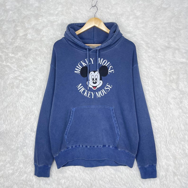 May include: A blue hooded sweatshirt featuring a Mickey Mouse graphic. The front of the sweatshirt displays the words "Mickey Mouse" in a circular design around a cartoon image of Mickey Mouse. The sweatshirt has a kangaroo pocket.