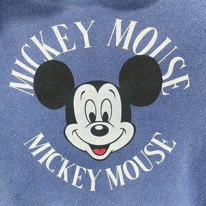May include: A blue hoodie featuring a large graphic of Mickey Mouse. The graphic includes Mickey's face, black ears, and the words "Mickey Mouse" in white, arched around the character. The hoodie has a front pocket and a drawstring hood.