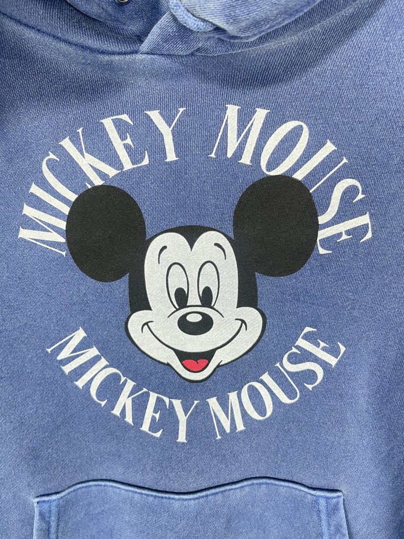 May include: A blue hoodie featuring a large graphic of Mickey Mouse. The graphic includes Mickey's face, black ears, and the words "Mickey Mouse" in white, arched around the character. The hoodie has a front pocket and a drawstring hood.