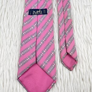 May include: A pink silk necktie featuring a repeating diagonal pattern of white and grey chain links. The back of the tie displays a black label with a carriage logo and the text 100% SILK, along with a white tag reading MADE IN FRANCE 5402 OA 100% SOIE.