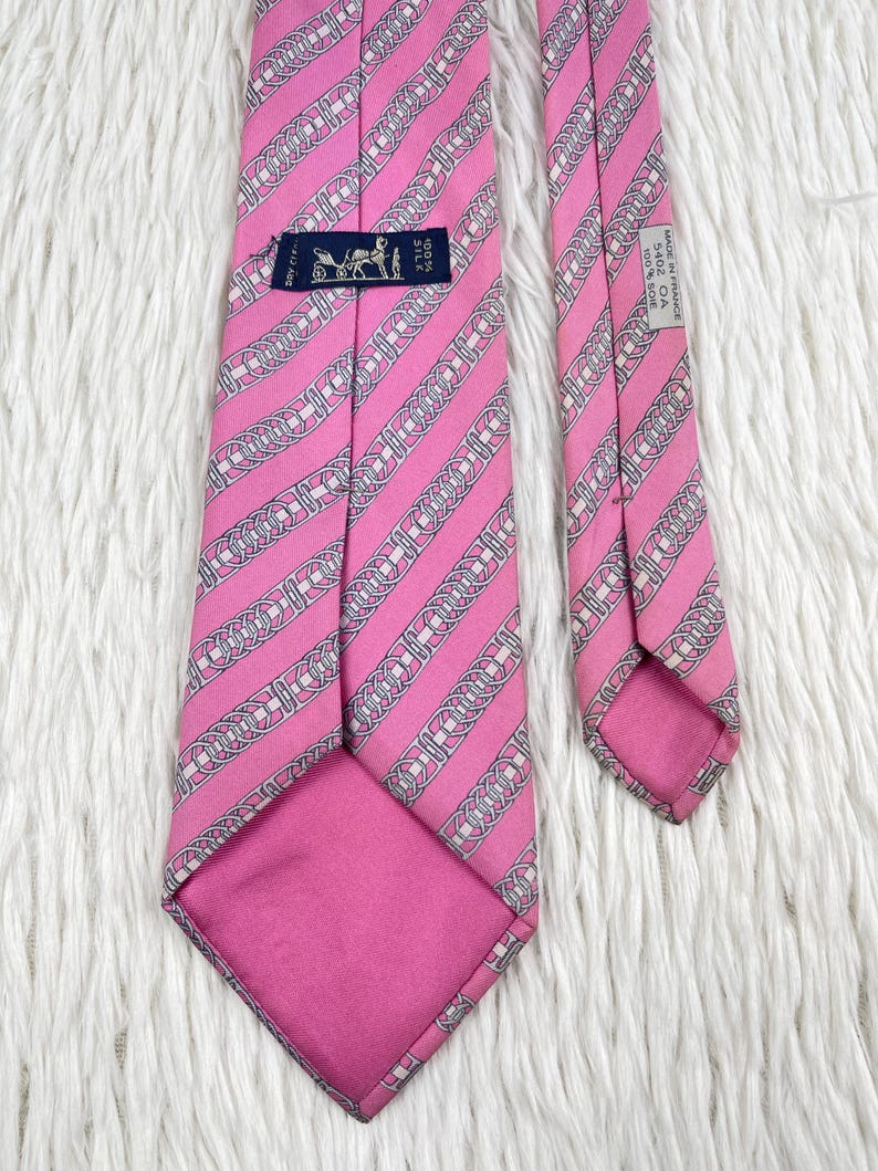 May include: A pink silk necktie featuring a repeating diagonal pattern of white and grey chain links. The back of the tie displays a black label with a carriage logo and the text 100% SILK, along with a white tag reading MADE IN FRANCE 5402 OA 100% SOIE.