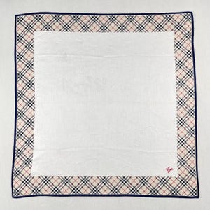 May include: Square white handkerchief with a beige, black, and red plaid border and a navy blue trim. The center is plain white. A small red logo is in the bottom right corner. The handkerchief is made of a soft, likely cotton, material.