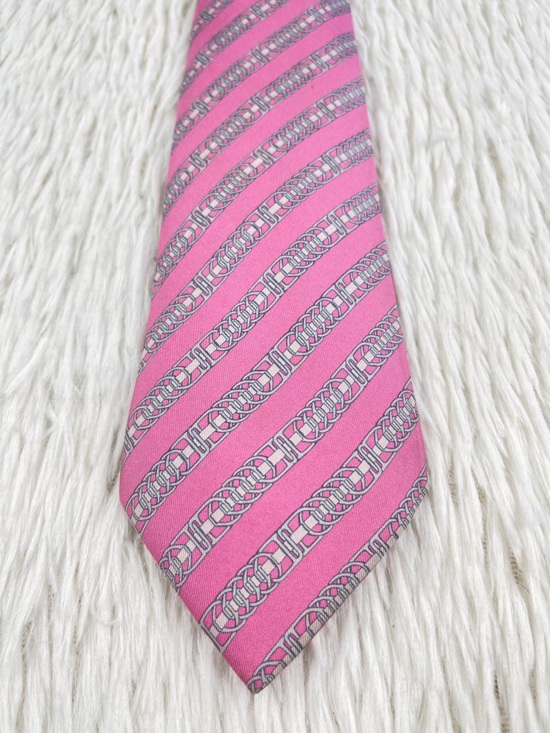 May include: A bright pink necktie featuring a repeating diagonal pattern of silver-toned chain links and interlocking circular motifs. The tie has a pointed tip and a smooth, uniform texture throughout the length of the fabric.