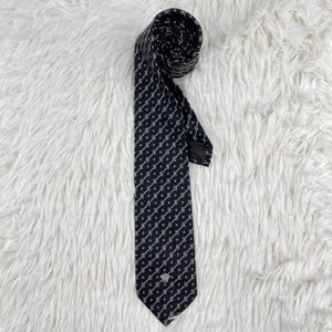 May include: A navy blue tie featuring a repeating white geometric design. The tie is rolled at the top and has a small emblem near the bottom. The tie is made of a smooth fabric.