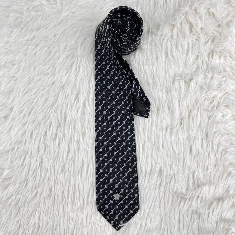 May include: A navy blue tie featuring a repeating white geometric design. The tie is rolled at the top and has a small emblem near the bottom. The tie is made of a smooth fabric.