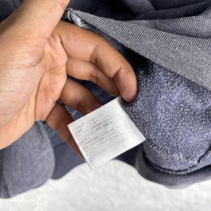 Może przedstawiać: Zbliżenie szarej bluzy z kapturem zapinanej na zamek. Bluza wykonana jest z miękkiej, teksturowanej tkaniny. Widoczna jest biała metka z japońskim tekstem. Zamek błyskawiczny jest w kolorze złotym.