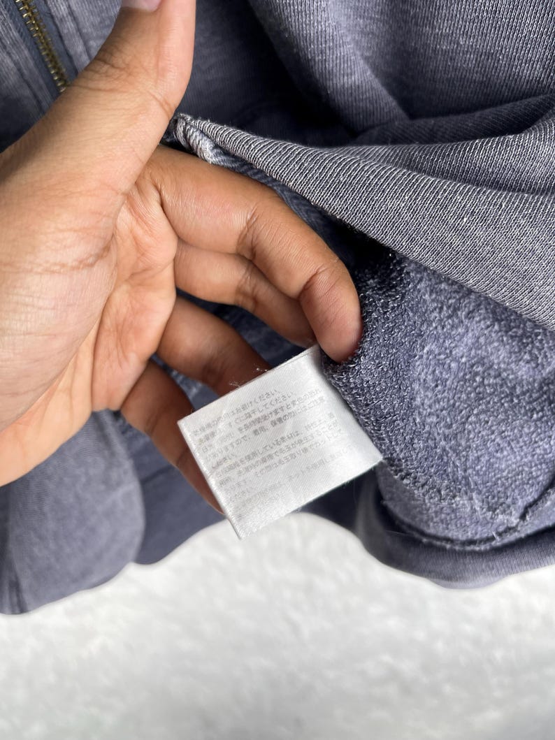 Może przedstawiać: Zbliżenie szarej bluzy z kapturem zapinanej na zamek. Bluza wykonana jest z miękkiej, teksturowanej tkaniny. Widoczna jest biała metka z japońskim tekstem. Zamek błyskawiczny jest w kolorze złotym.