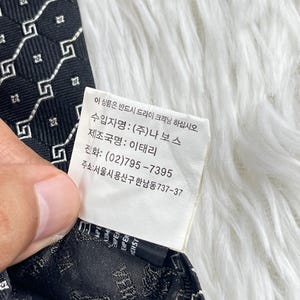 May include: A black tie featuring a repeating white geometric design. The label includes Korean text and details about the Italian manufacturer.