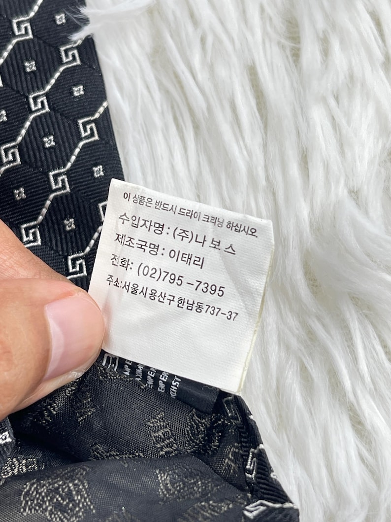 May include: A black tie featuring a repeating white geometric design. The label includes Korean text and details about the Italian manufacturer.