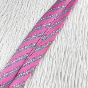 May include: A bright pink necktie featuring a repeating pattern of silver-toned chain links and horizontal stripes. The fabric has a smooth, satin-like finish with diagonal lines running across the length of the tie, creating a structured appearance.