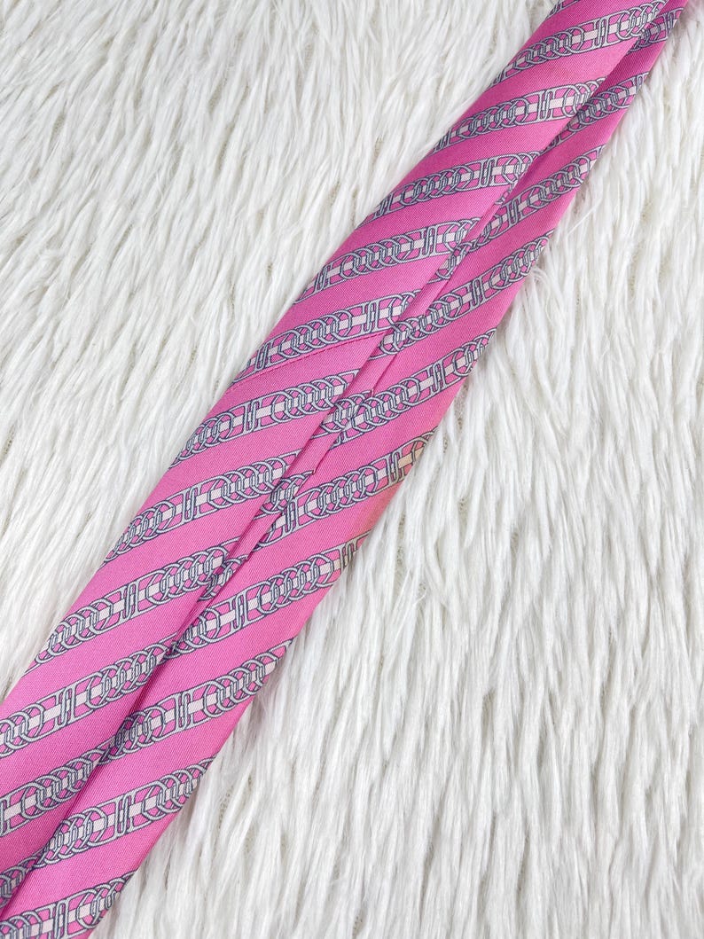 May include: A bright pink necktie featuring a repeating pattern of silver-toned chain links and horizontal stripes. The fabric has a smooth, satin-like finish with diagonal lines running across the length of the tie, creating a structured appearance.
