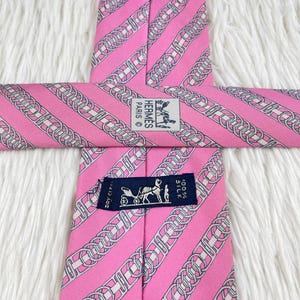 May include: A pink silk necktie featuring a repeating pattern of diagonal silver and white chain links. The tie includes a white label with the text HERMES PARIS and a dark blue label with a horse-drawn carriage graphic and the text 100% SILK.