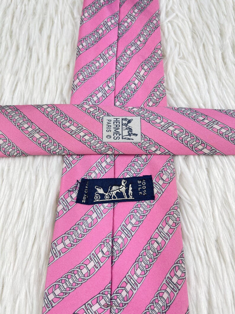 May include: A pink silk necktie featuring a repeating pattern of diagonal silver and white chain links. The tie includes a white label with the text HERMES PARIS and a dark blue label with a horse-drawn carriage graphic and the text 100% SILK.