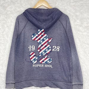 Może przedstawiać: Niebieska bluza z kapturem z nadrukiem Myszki Miki na plecach. Sylwetka Myszki Miki wypełniona jest czerwono-białymi paskami i niebieskimi gwiazdami. Pod spodem znajduje się napis "19 28" i "SUPER IDOL".