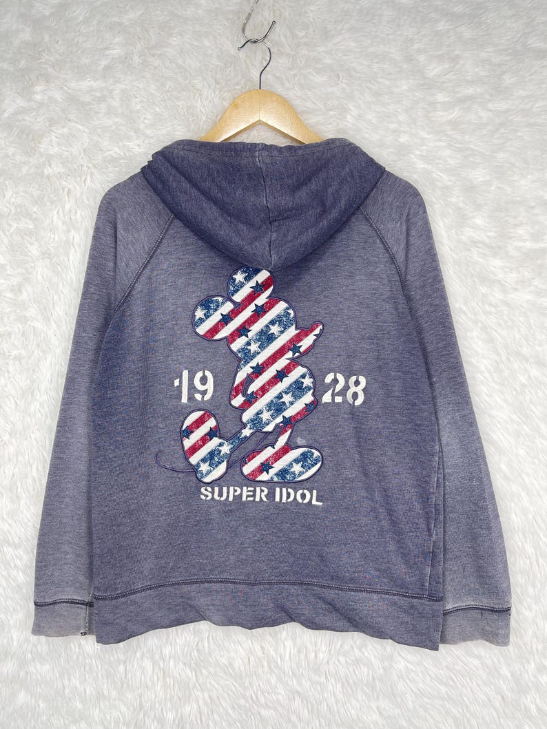 Może przedstawiać: Niebieska bluza z kapturem z nadrukiem Myszki Miki na plecach. Sylwetka Myszki Miki wypełniona jest czerwono-białymi paskami i niebieskimi gwiazdami. Pod spodem znajduje się napis "19 28" i "SUPER IDOL".