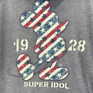 Może przedstawiać: Szara bluza z kapturem z nadrukiem Myszki Miki w czerwono-biało-niebieskie paski i gwiazdy. Pod postacią nadrukowano napis "1928 SUPER IDOL". Bluza ma kaptur ze sznurkiem.