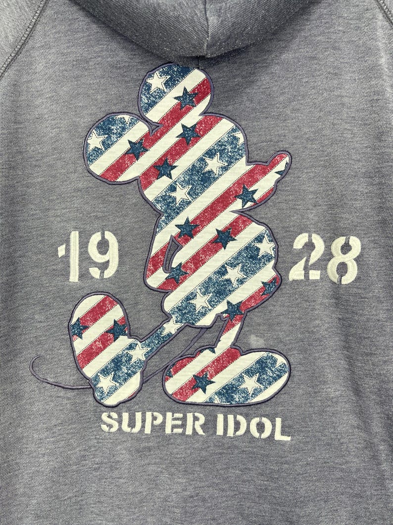 Może przedstawiać: Szara bluza z kapturem z nadrukiem Myszki Miki w czerwono-biało-niebieskie paski i gwiazdy. Pod postacią nadrukowano napis "1928 SUPER IDOL". Bluza ma kaptur ze sznurkiem.
