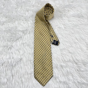 Puede incluir: Una corbata estampada en amarillo y negro. La corbata está enrollada en la parte superior y se encuentra en diagonal sobre una superficie blanca texturizada. El patrón consiste en pequeñas formas geométricas repetidas en amarillo y negro.