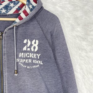 Może przedstawiać: Szara bluza z kapturem zapinana na zamek z kapturem wyłożonym czerwono-biało-niebieskim wzorem w gwiazdy i paski. Prz&oacute;d bluzy z kapturem zdobi liczba "28" oraz białe napisy "Mickey Super Idol Great Big Heart".