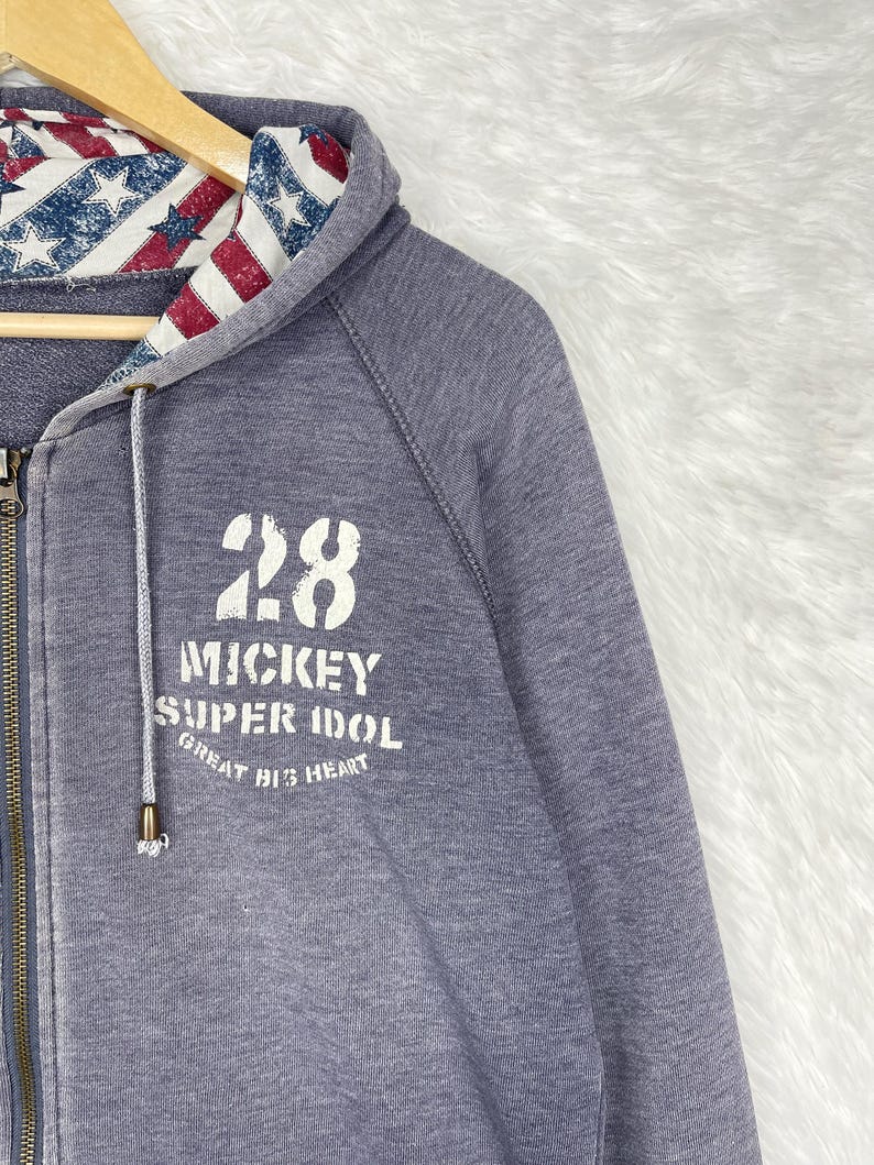 Może przedstawiać: Szara bluza z kapturem zapinana na zamek z kapturem wyłożonym czerwono-biało-niebieskim wzorem w gwiazdy i paski. Prz&oacute;d bluzy z kapturem zdobi liczba "28" oraz białe napisy "Mickey Super Idol Great Big Heart".