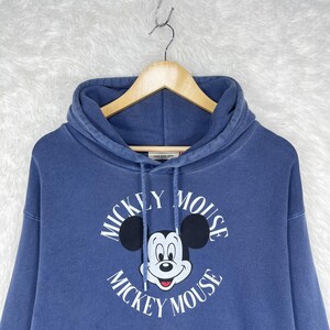 May include: A blue hooded sweatshirt featuring a Mickey Mouse graphic. The graphic includes Mickey's face and the words "Mickey Mouse" in a circular design. The sweatshirt has a front pocket and is hanging on a wooden hanger.