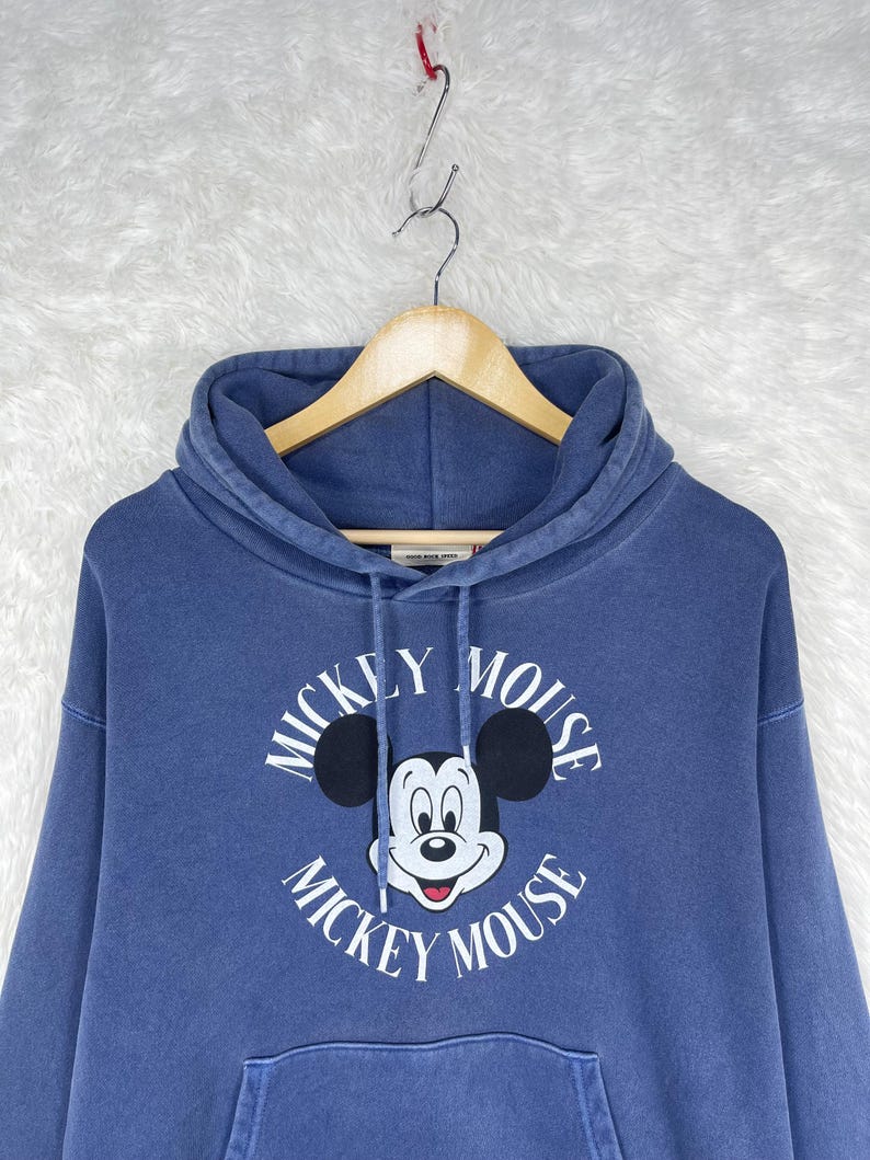 May include: A blue hooded sweatshirt featuring a Mickey Mouse graphic. The graphic includes Mickey's face and the words "Mickey Mouse" in a circular design. The sweatshirt has a front pocket and is hanging on a wooden hanger.