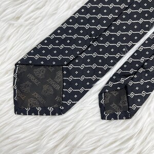 May include: A black tie with a repeating white geometric pattern. The tie's interior lining features the Versace logo. The tie is displayed on a white, fluffy surface.