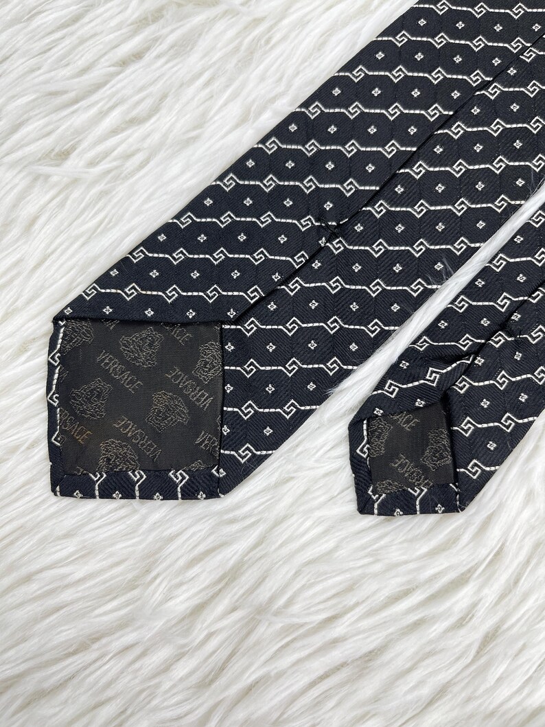 May include: A black tie with a repeating white geometric pattern. The tie's interior lining features the Versace logo. The tie is displayed on a white, fluffy surface.