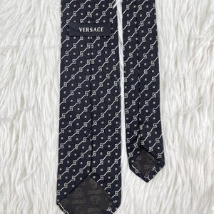 May include: A black Versace tie with a repeating white geometric pattern. The tie is made of a smooth fabric and features the Versace logo on the label and the tip.