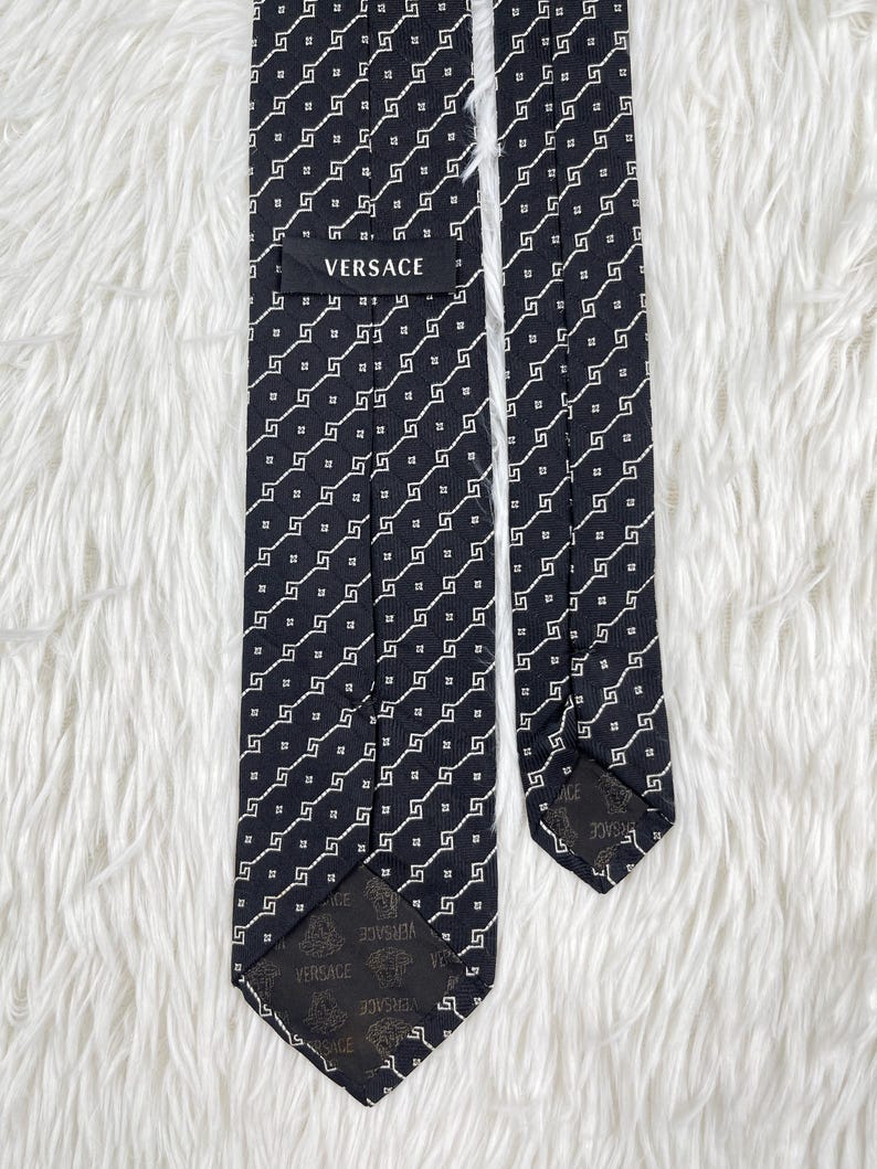May include: A black Versace tie with a repeating white geometric pattern. The tie is made of a smooth fabric and features the Versace logo on the label and the tip.