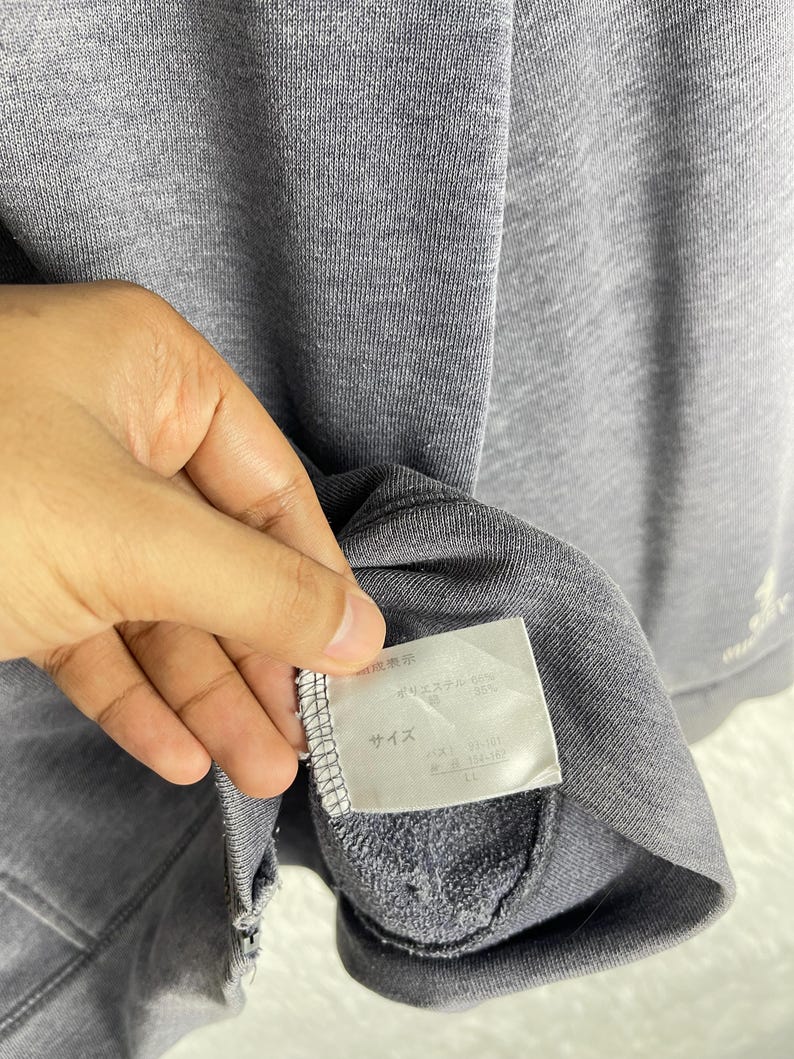 Może przedstawiać: Szara bluza z dołączoną metką. Etykieta zawiera japoński tekst i szczeg&oacute;ły. Bluza wykonana jest z miękkiej, teksturowanej tkaniny. Rękaw jest złożony, odsłaniając wnętrze odzieży.