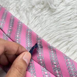 May include: A pink necktie featuring a repeating pattern of vertical silver and white chain-link motifs. The fabric has a subtle diagonal ribbed texture, and a small dark blue fabric label is attached to the back of the tie.