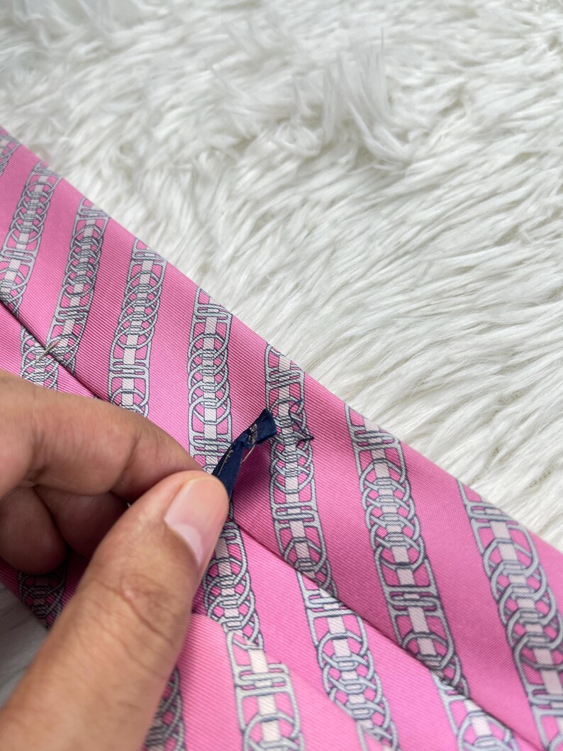 May include: A pink necktie featuring a repeating pattern of vertical silver and white chain-link motifs. The fabric has a subtle diagonal ribbed texture, and a small dark blue fabric label is attached to the back of the tie.