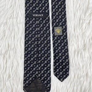 May include: A black Versace tie with a repeating white geometric pattern. The tie features the Versace logo on a label and a gold-coloured emblem with a Medusa head. The tie is displayed on a white, textured surface.