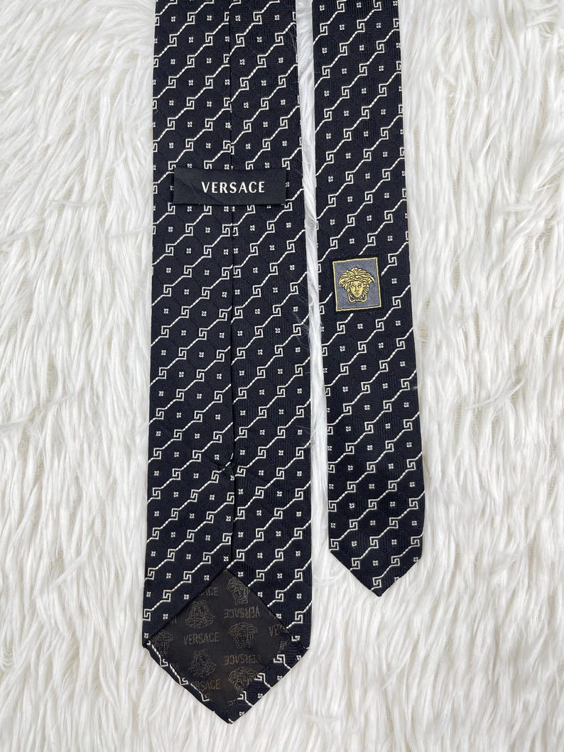 May include: A black Versace tie with a repeating white geometric pattern. The tie features the Versace logo on a label and a gold-coloured emblem with a Medusa head. The tie is displayed on a white, textured surface.