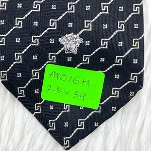 May include: Black necktie with a repeating white geometric pattern and a Medusa head emblem. The tie has a green tag with the text "ATO16H" and measurements of 6.35 x 149.86 cm.