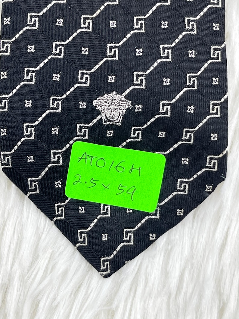 May include: Black necktie with a repeating white geometric pattern and a Medusa head emblem. The tie has a green tag with the text "ATO16H" and measurements of 6.35 x 149.86 cm.