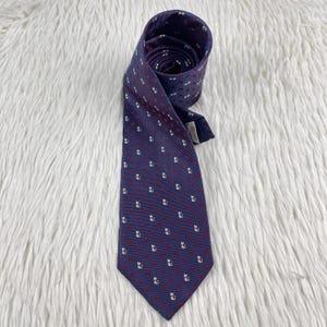 Puede incluir: Una corbata azul oscuro con un patrón repetitivo de pequeñas siluetas de gatos blancas y diminutos puntos rojos. La corbata está enrollada en la parte superior, mostrando una superficie de tela texturizada con un extremo puntiagudo y etiqueta.