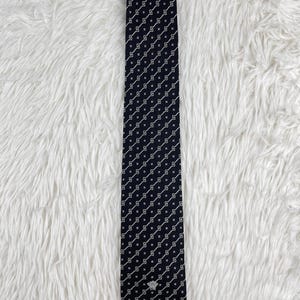 May include: A navy blue tie with a repeating white geometric pattern. The tie has a small silver emblem near the pointed end. The tie is displayed on a white, textured surface.