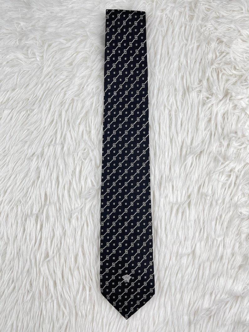 May include: A navy blue tie with a repeating white geometric pattern. The tie has a small silver emblem near the pointed end. The tie is displayed on a white, textured surface.