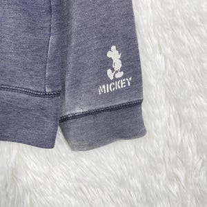 Może przedstawiać: Zbliżenie na wyblakły niebieski rękaw bluzy. Biała grafika Myszki Miki i napis "MICKEY" są nadrukowane na mankiecie. Tkanina ma lekko zużyty wygląd.