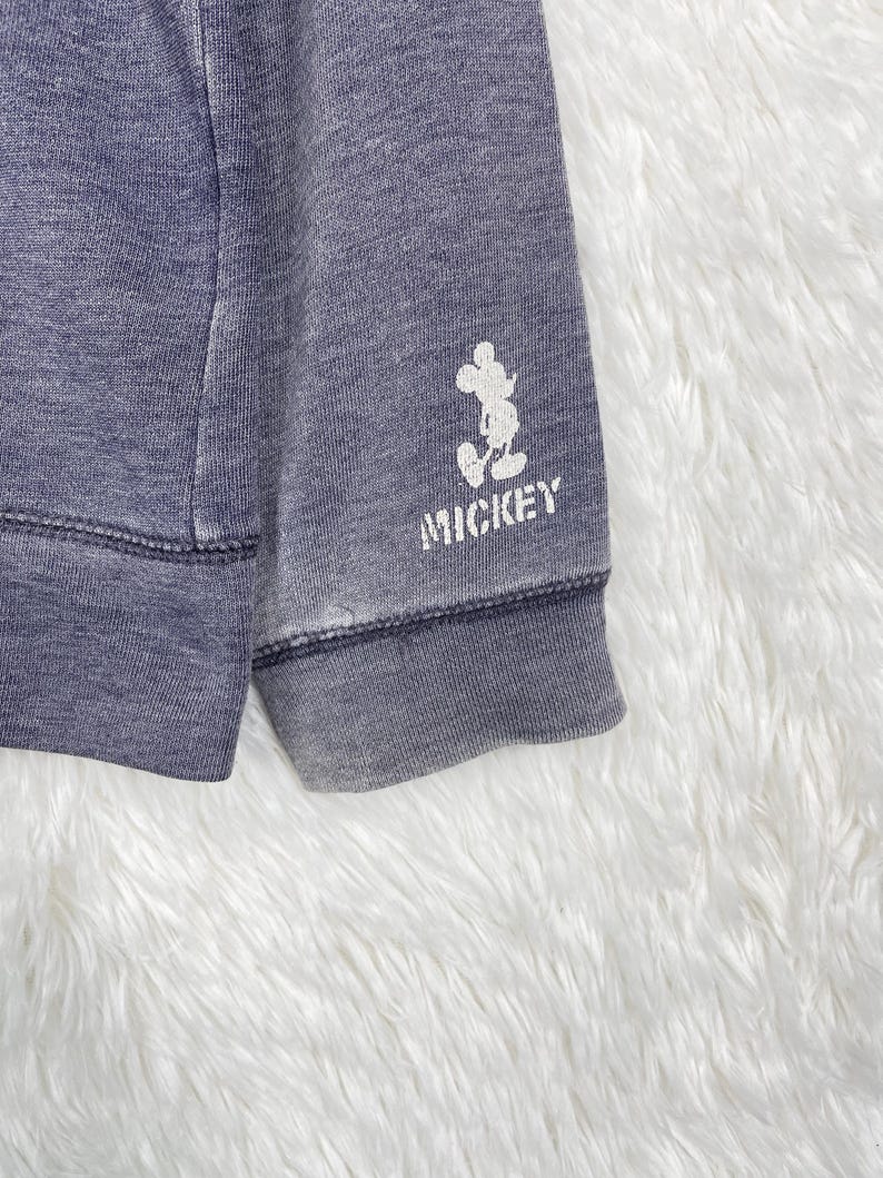 Może przedstawiać: Zbliżenie na wyblakły niebieski rękaw bluzy. Biała grafika Myszki Miki i napis "MICKEY" są nadrukowane na mankiecie. Tkanina ma lekko zużyty wygląd.