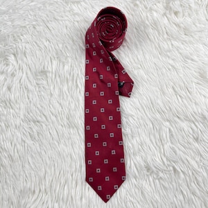 Puede incluir: Una corbata burdeos enrollada con un patrón repetido de pequeños cuadrados blancos. La corbata está hecha de una tela suave y se muestra sobre una superficie blanca y esponjosa. Es un accesorio clásico.